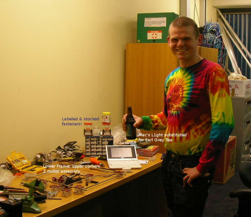 A picture of Sam holding a beer standing by a well stocked set of fastenings and a RepRap in early stages of construction